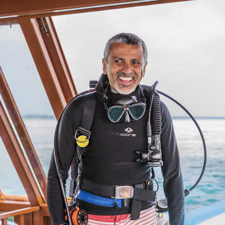 Luiz Rocha stands atop a boat in the Maldives wearing a black scuba diving outfit, a rebreather and goggles, and trunks.