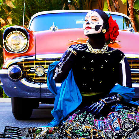 Woman with calavera face paint in traditional dress sitting on ground in front of old red Chevy