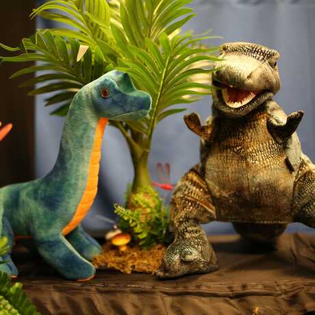 A sauropod and Allosaurus puppet in a prehistoric puppet theater landscape