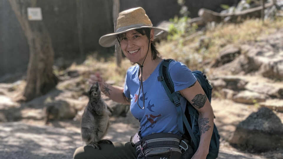 Sarah Crews feeds wallabies in Australia while searching for flattie spiders. She wears a blue shirt and hiking gear. 
